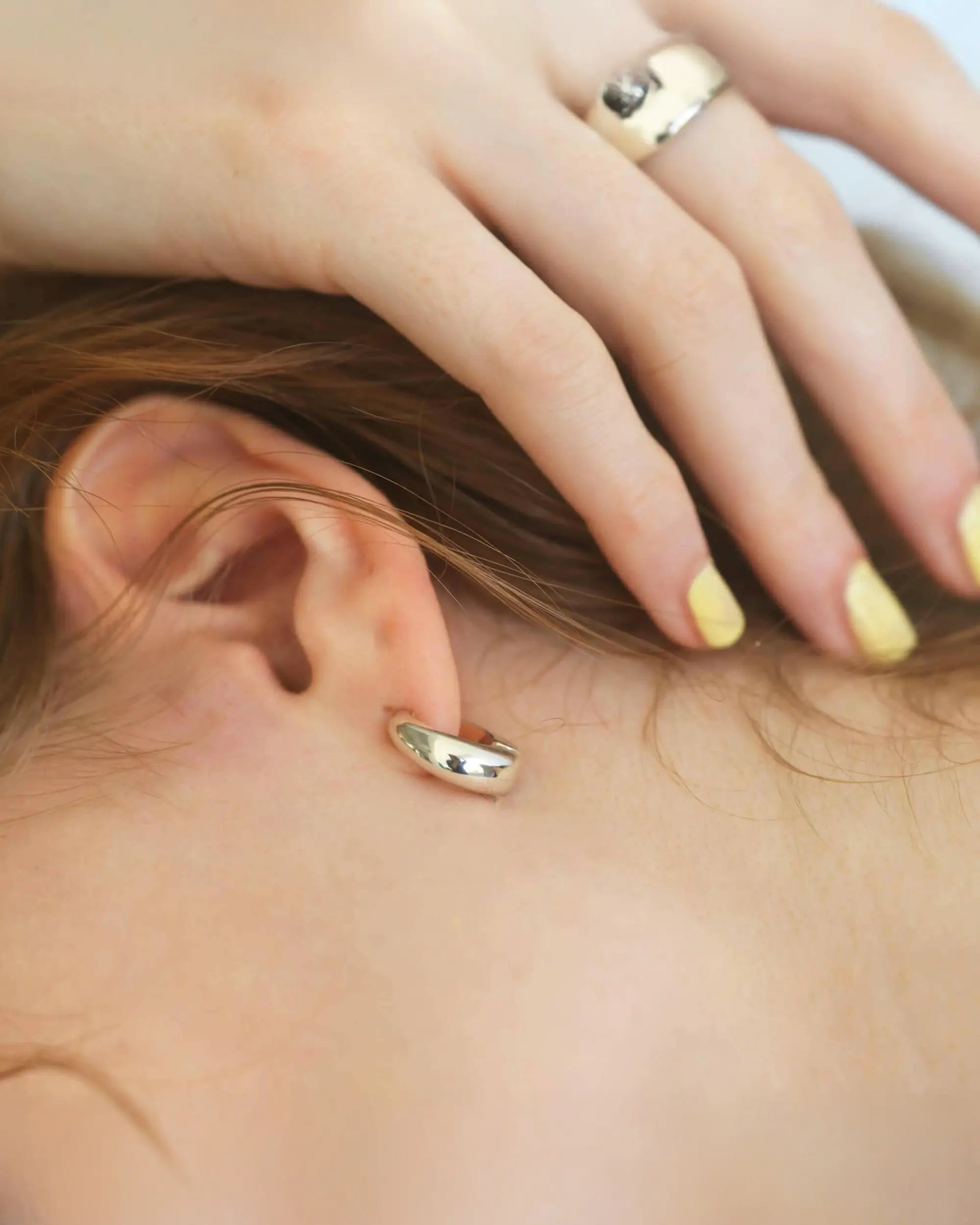 耳への負担が少ないバネ式構造のシルバーノンホールピアスを着用した、女性の耳元と首筋の美しいクローズアップ画像。イエローのネイルがアクセント。