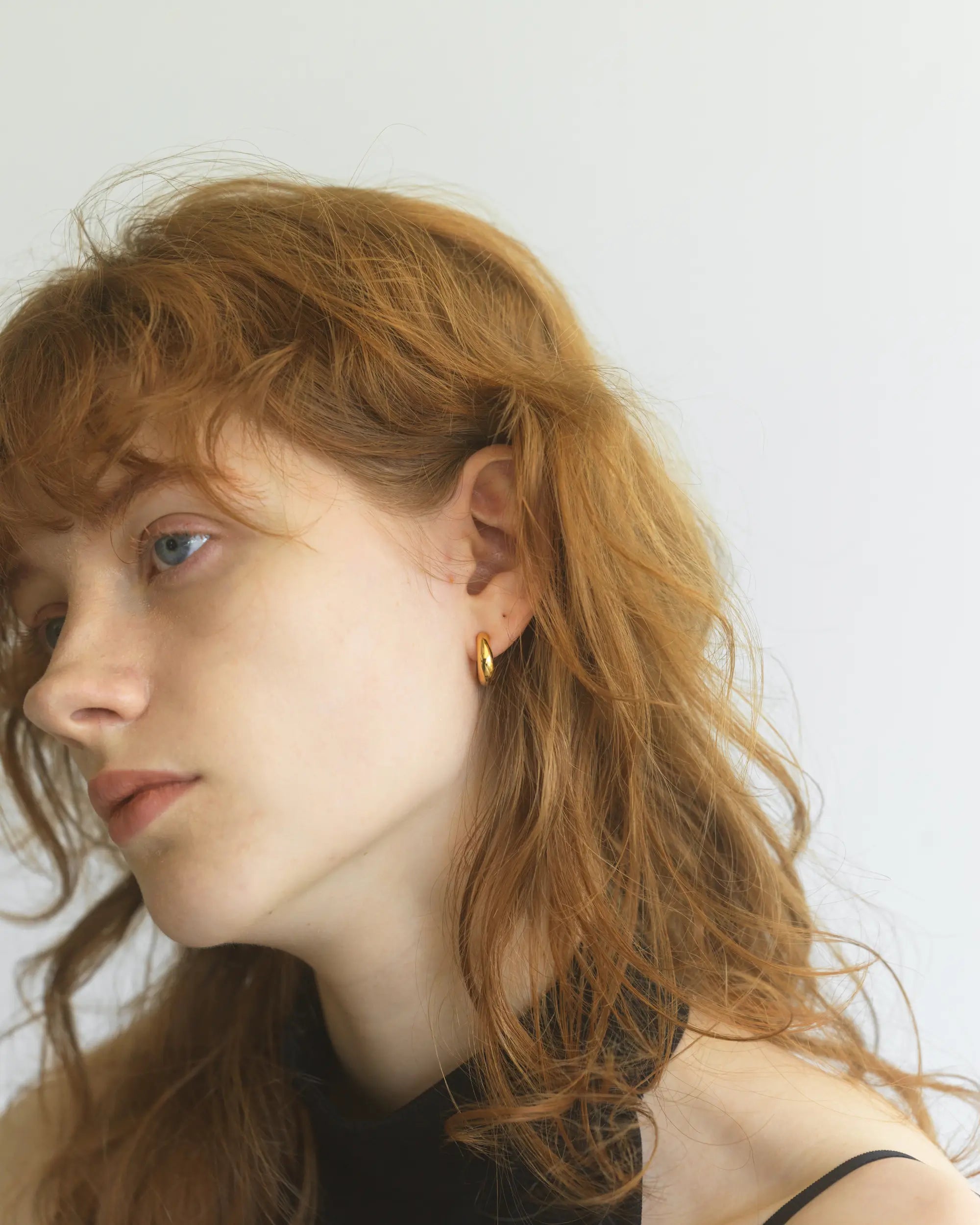 黒いトップスを着た女性が、厚みのあるゴールドの大ぶりフープイヤリングを着用して斜め下を見つめるアンニュイな横顔。