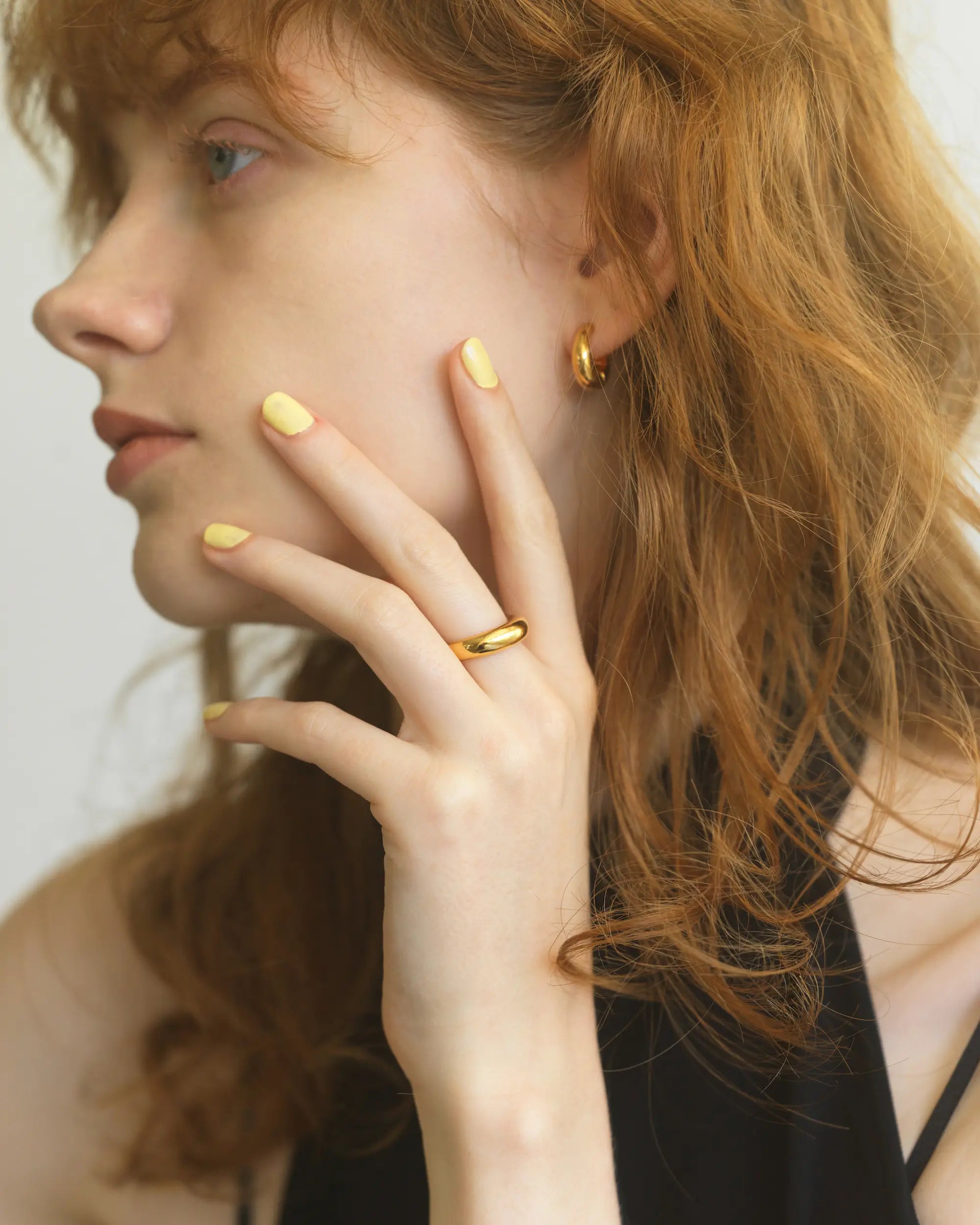 イエローのネイルを施した手を顔に添え、18Kゴールドコーティングのチャンキーなフープイヤリングを着用した女性の横顔ポートレート。