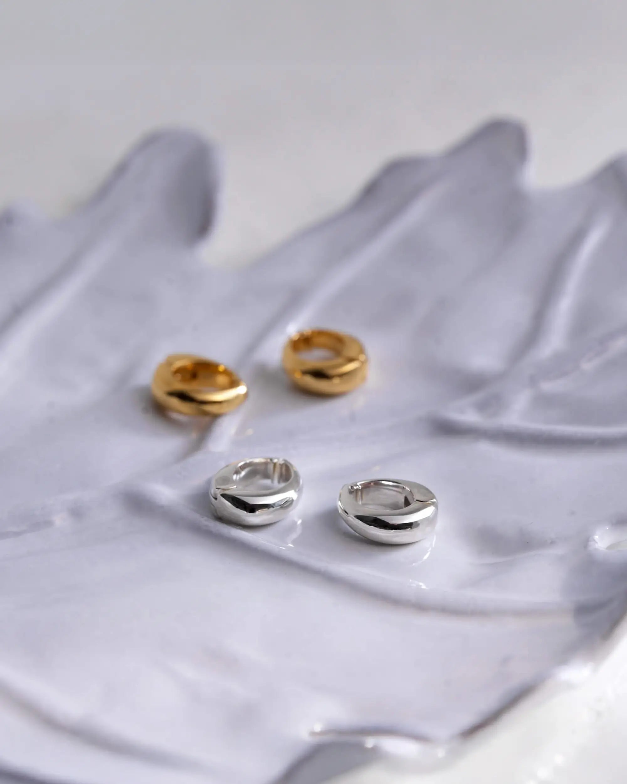 グレーの波打つアートオブジェの上に置かれた、シルバーとゴールドのバネ式ヒンジドフープイヤリング(大ぶり)のエモーショナルな置き画。