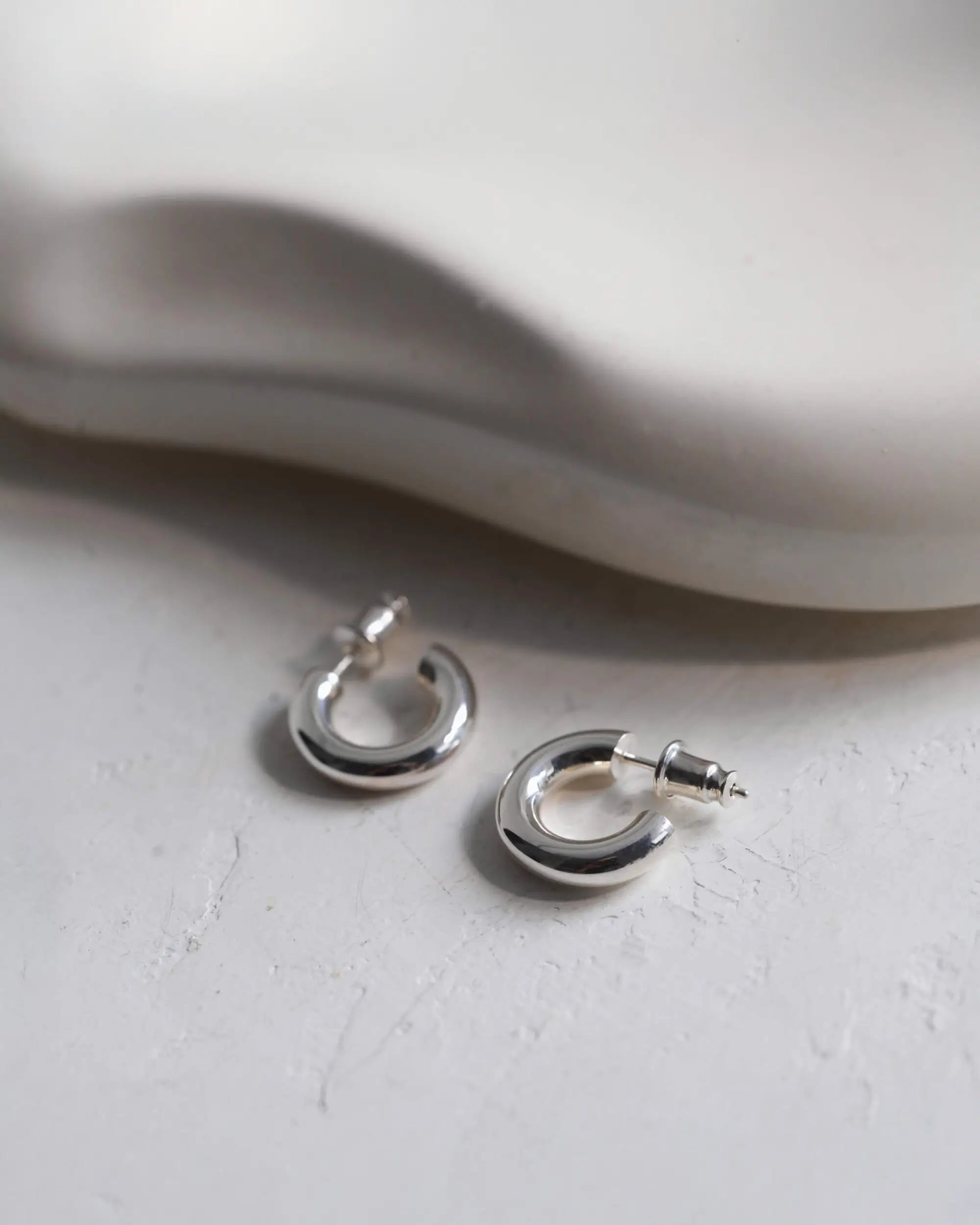 白いストーンの上に置かれたオーバルフープピアス S(シルバー)の置き画。クラシックな美しさを現代的に解釈した、大人のためのタイムレスなスキンジュエリー。