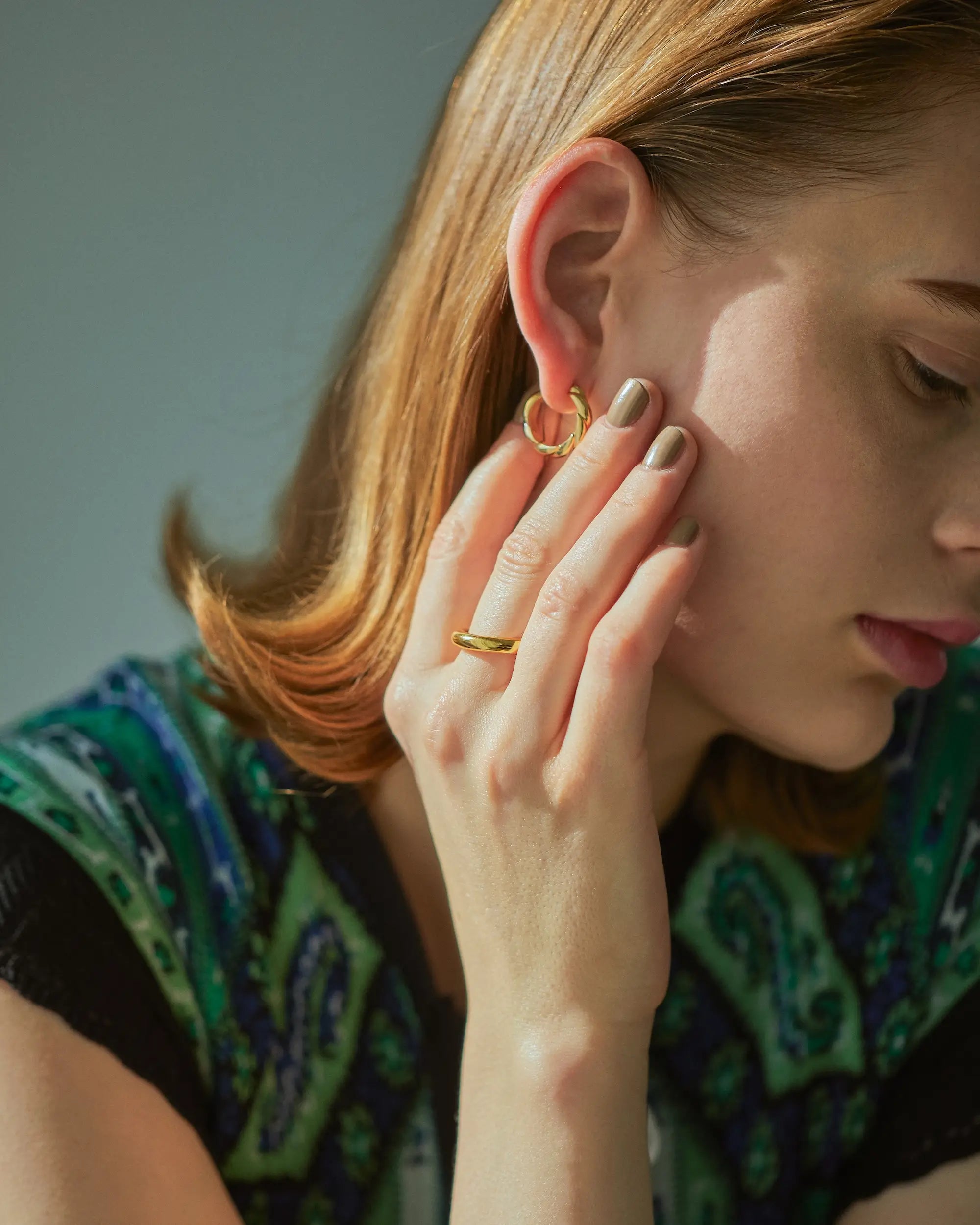 柄物のトップスを着た女性が、18Kゴールドコーティングの大ぶりなウェーブピアスを着用して、リングを着けた手を顔に添えているエレガントな横顔。