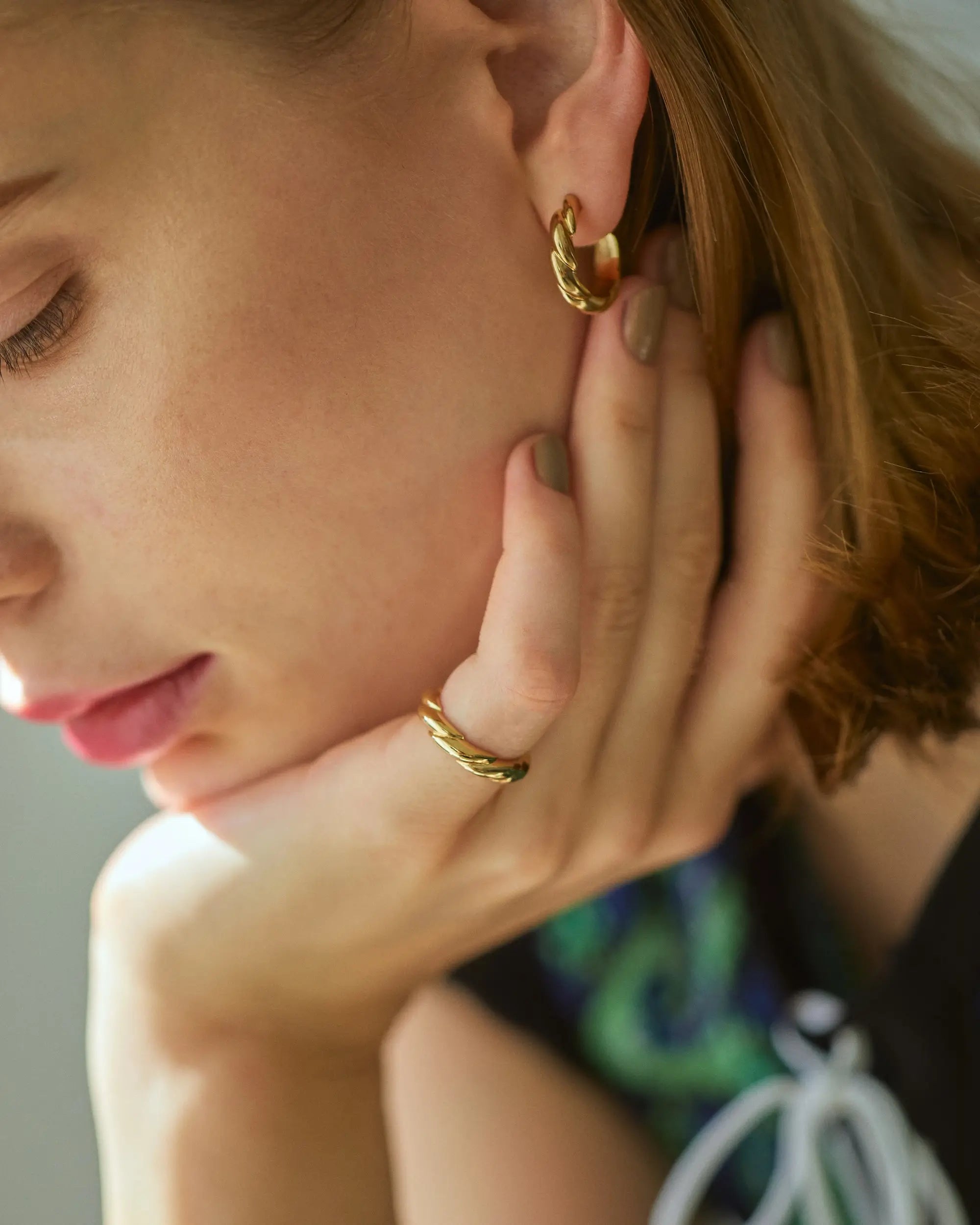 リングを着けた手を首元に添えてうつむく女性モデルの耳元で、華やかな存在感を放つ大ぶりなゴールドのウェーブピアス。
