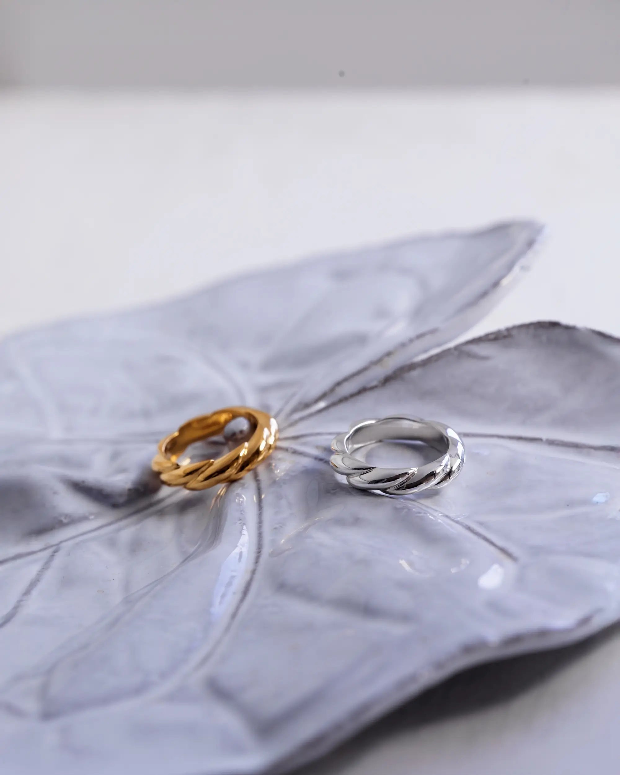 グレーの陶器に置かれたウェーブリング(ゴールドとシルバー)のペア置き画。大切な方へのギフトやペアリングにもおすすめの曲線美。