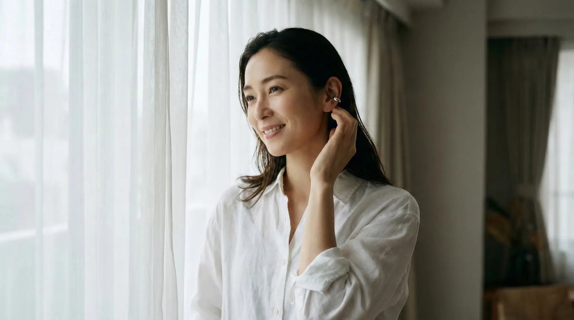 窓辺でリラックスしながらシルバーのイヤーカフを身につける30代女性