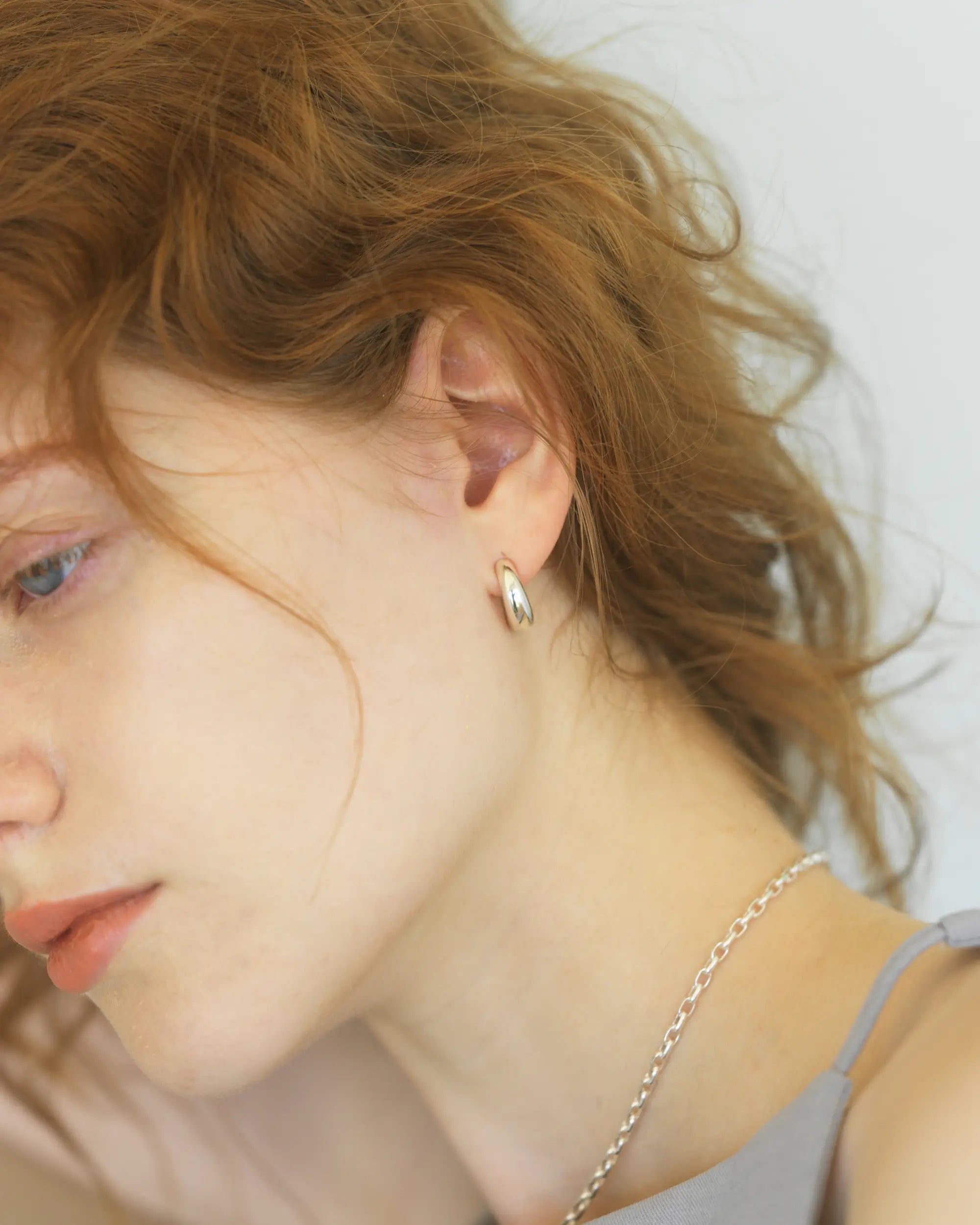 グレーのキャミソールを着た女性が、金具が見えない「ピアス見え」するシルバーのヒンジドフープイヤリング（Mサイズ）を着用した、洗練された横顔のポートレート。