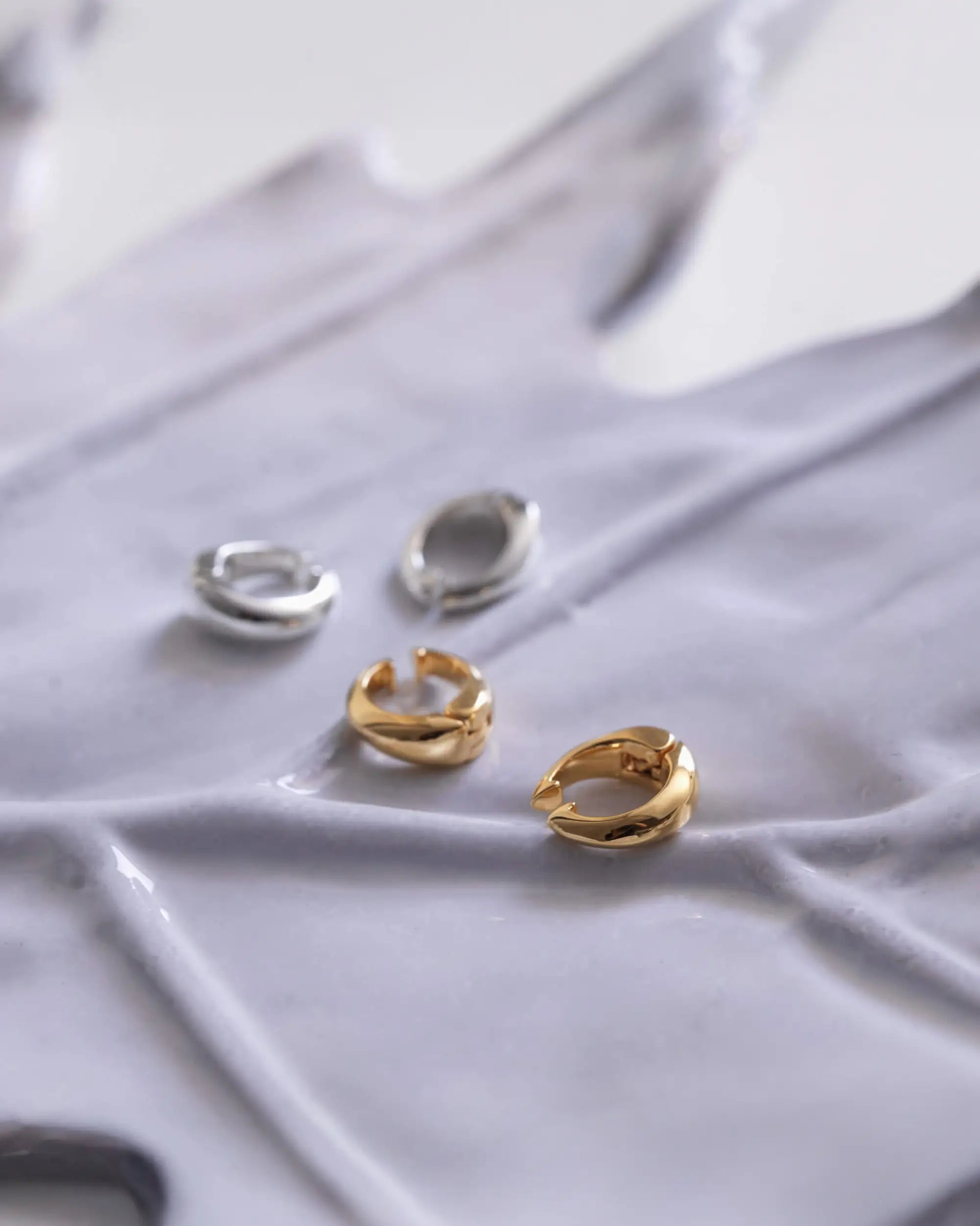 波打つグレーのオブジェの上に置かれた、シルバー925製とゴールドのヒンジドフープイヤリング（Sサイズ）。イヤーカフとしても使える有機的なフォルムの置き画。