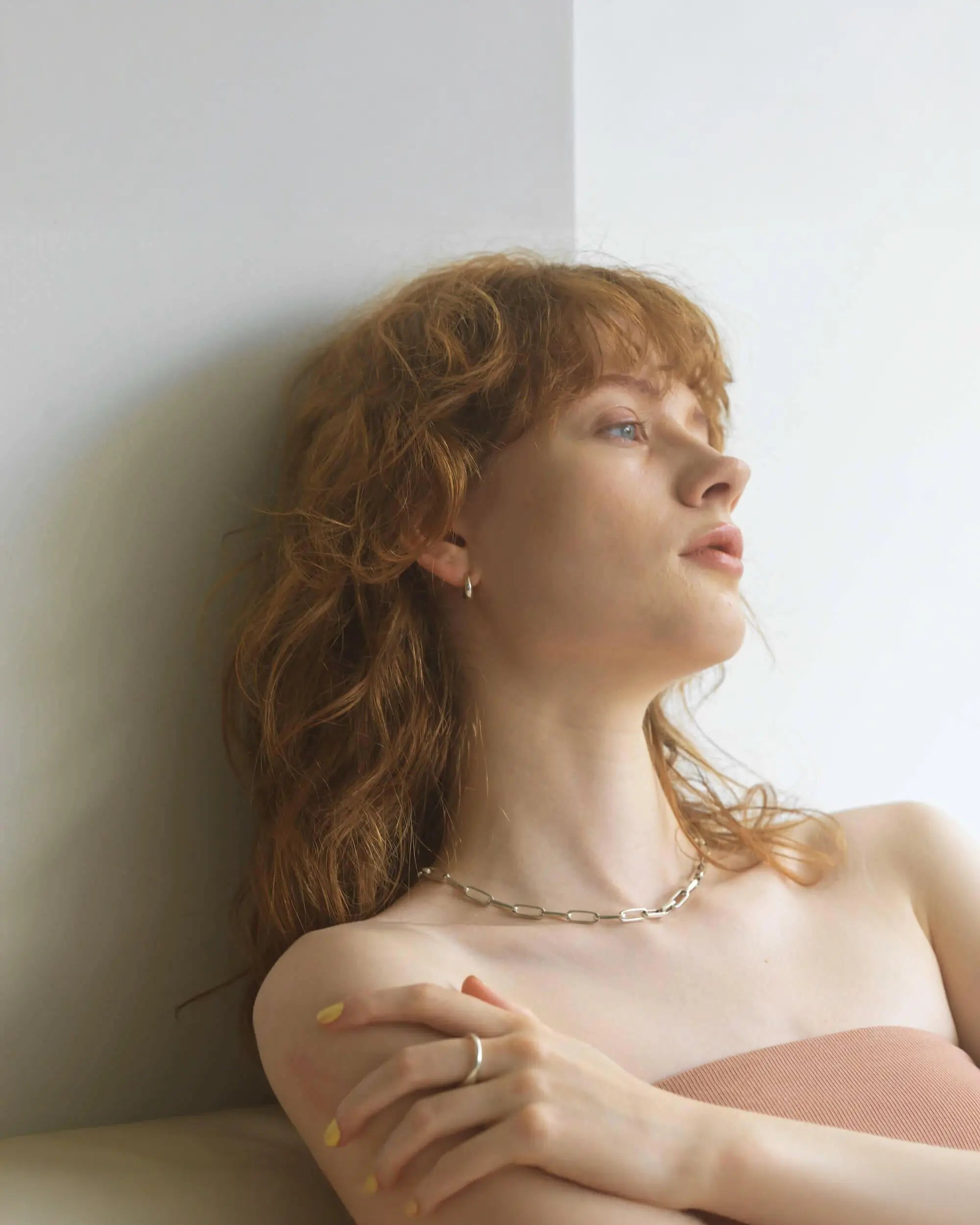 白い壁にもたれる、赤毛の女性モデルのエフォートレスなポートレート。ピアスのように見えるフープイヤリングを着用しています。