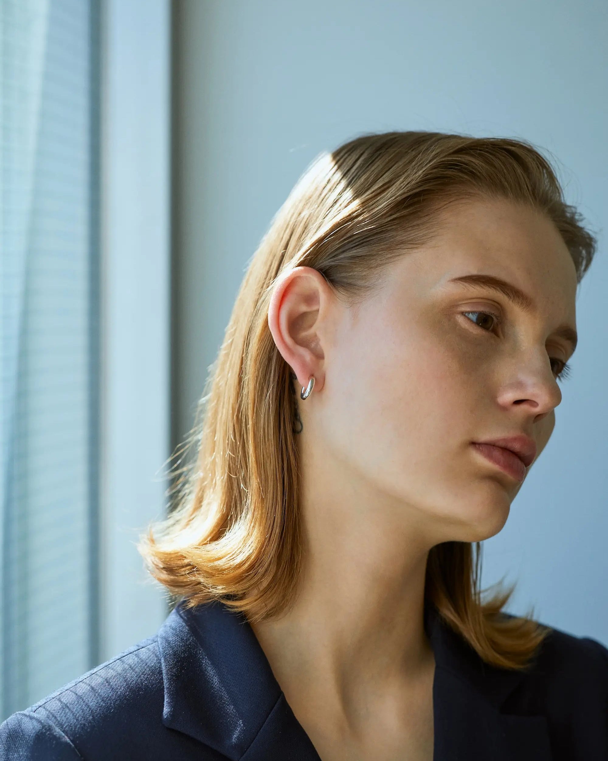 シルバー925製オーバルフープピアス Sを着用した女性モデル。ネイビージャケットなどオフィスでのビジネススタイルにも上品に馴染む知的で小ぶりなデザイン。