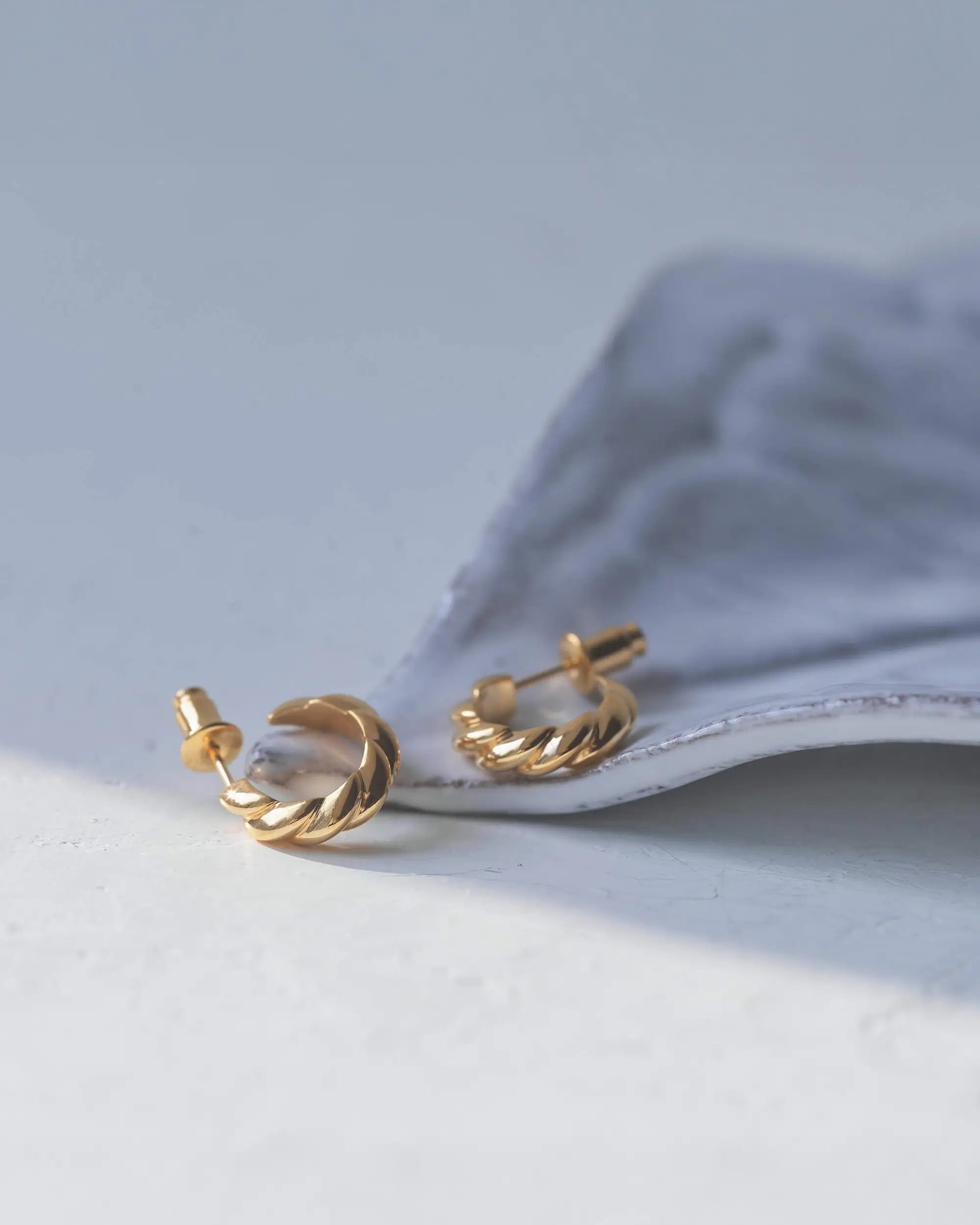グレーの布地の上に置かれた、18Kゴールドコーティングが美しいウェーブピアス（Sサイズ）。海面の波をモチーフにした立体的で柔らかな曲線が光を反射しています。